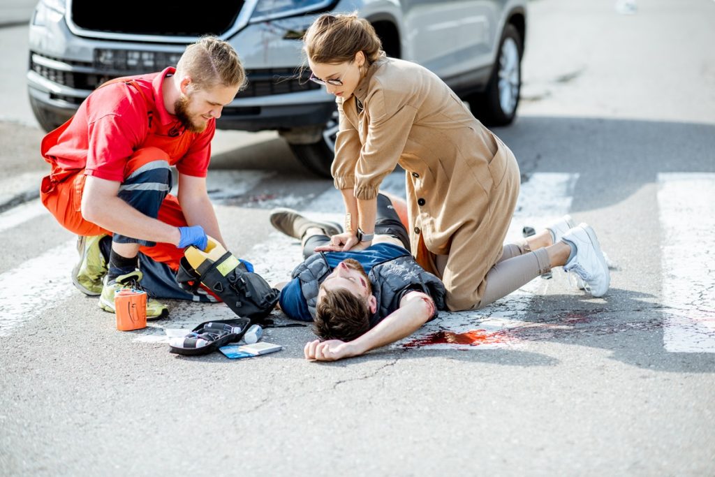 Two people helping an injured man lying in the road after a car accident